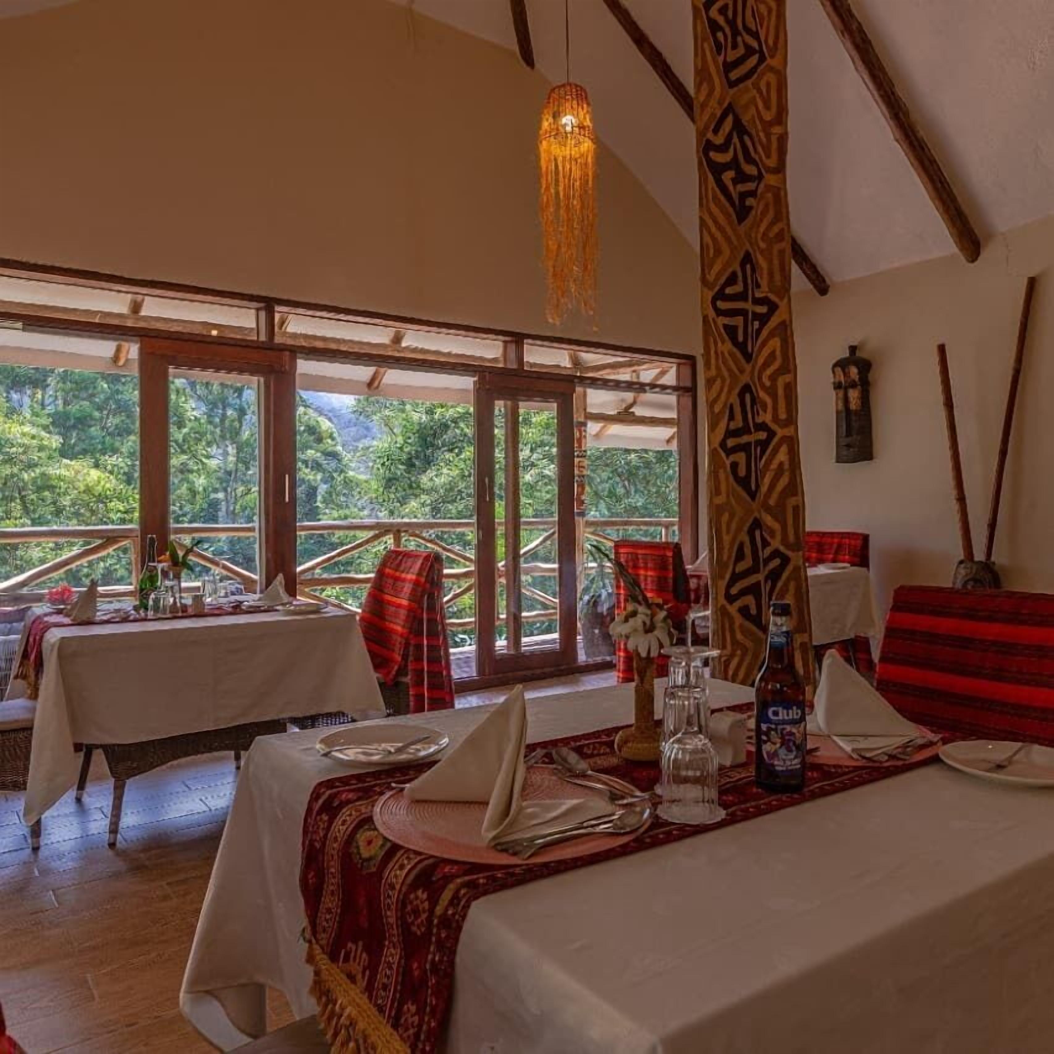 A dining area with tables set for two, a hanging light fixture, and a view of greenery outside.
