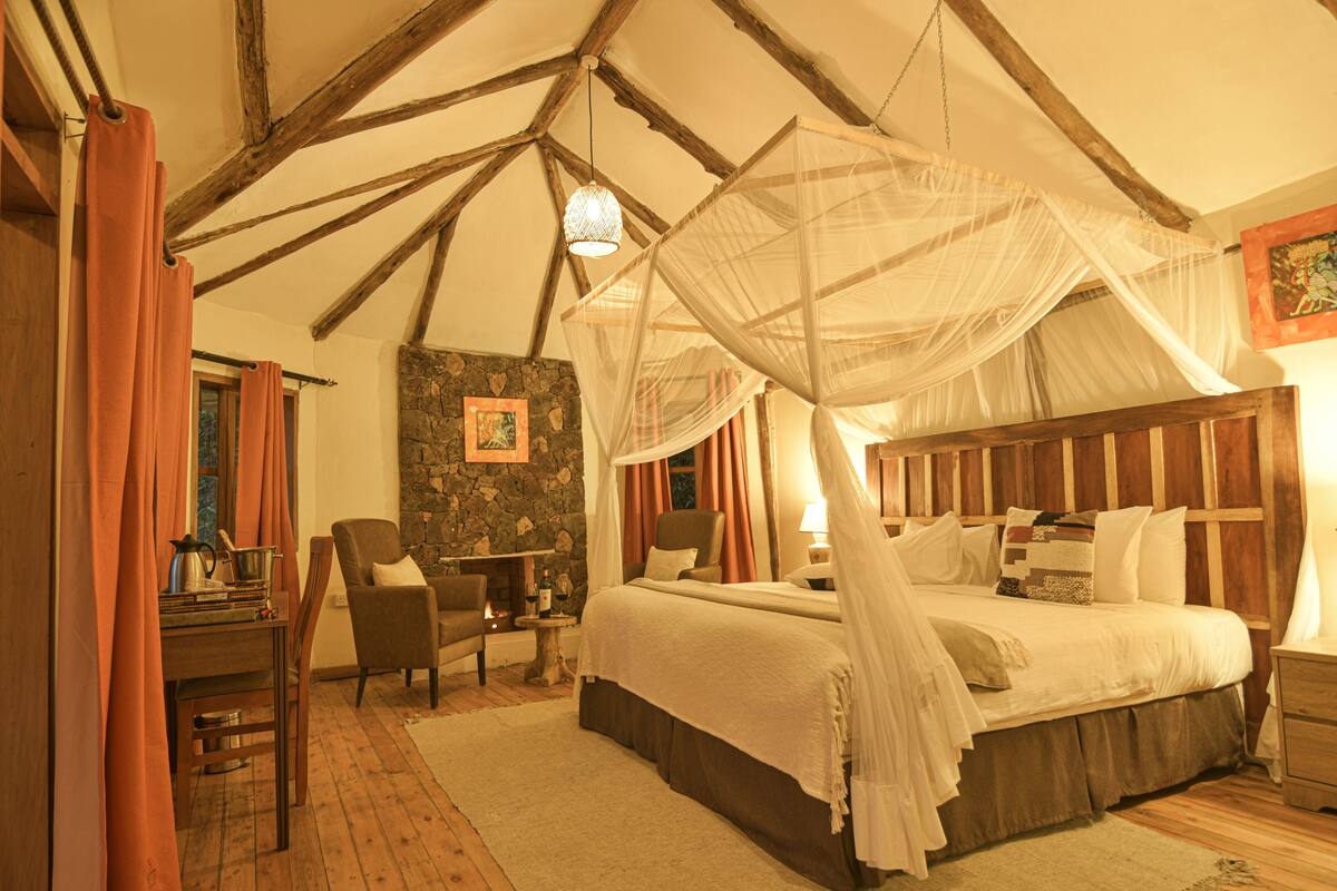 A bedroom with a four-poster bed, a stone wall, a wooden ceiling, and a small table with a lamp.