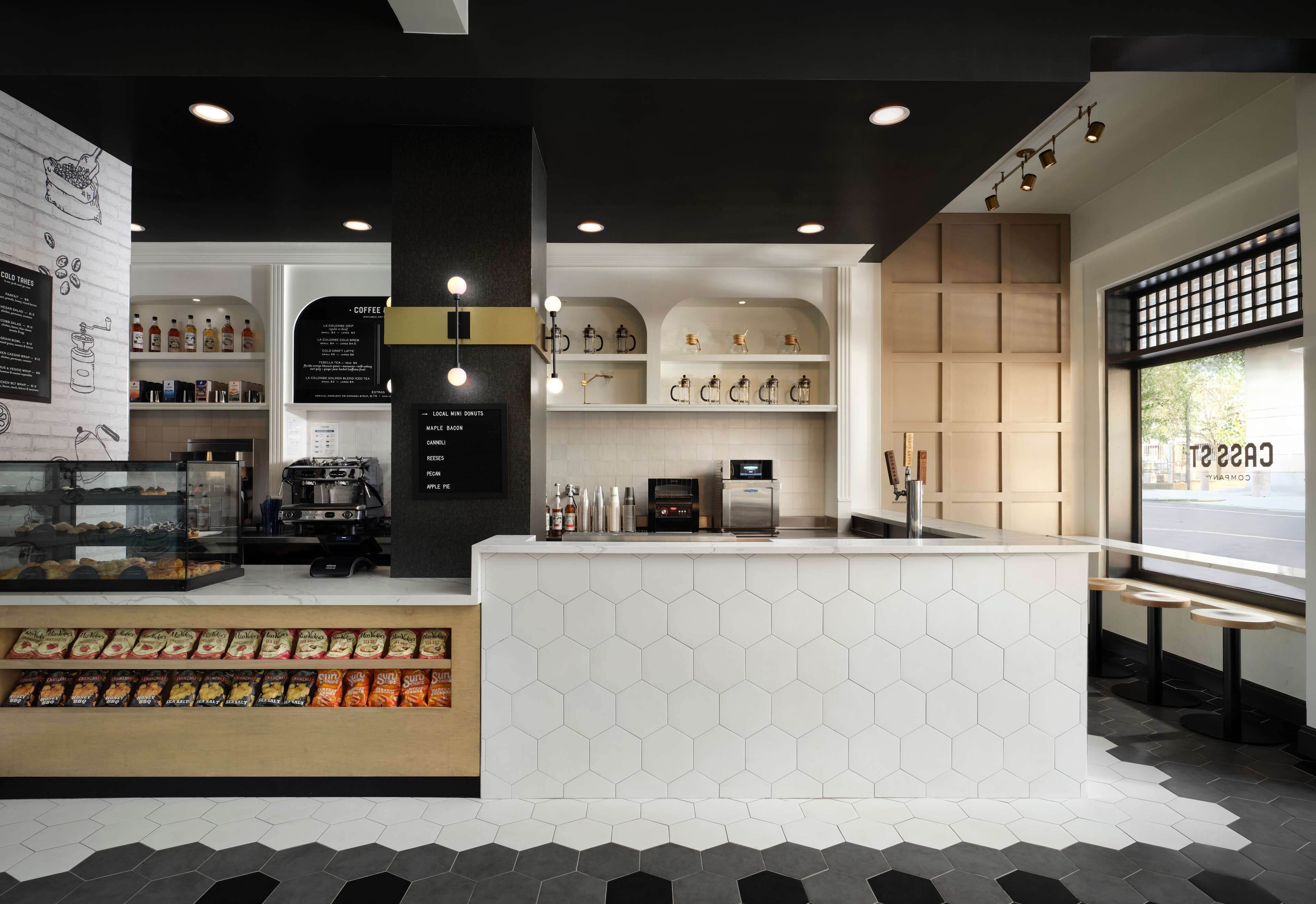 Un café moderne avec un comptoir recouvert de carreaux hexagonaux, des étagères remplies de produits à base de café et de thé, et un présentoir de collations.