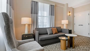 A hotel room with a grey sofa, two bedside lamps, a patterned armchair, a small round table, and a patterned carpet.