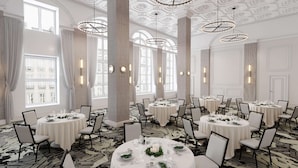 Een ruime eetruimte met meerdere ronde tafels, witte tafelkleden en moderne kroonluchters.