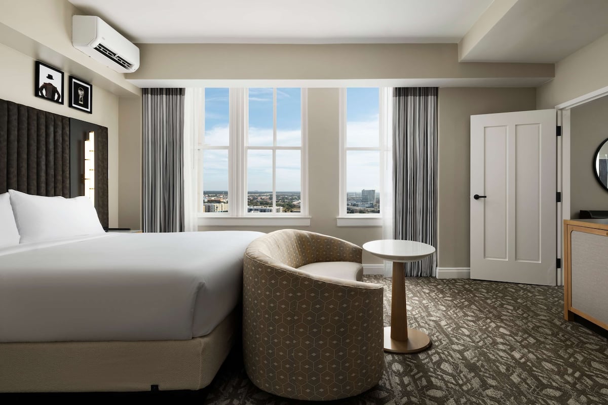 Een hotelkamer met een groot bed, een gebogen fauteuil, een kleine ronde tafel en uitzicht op de stad door grote ramen.