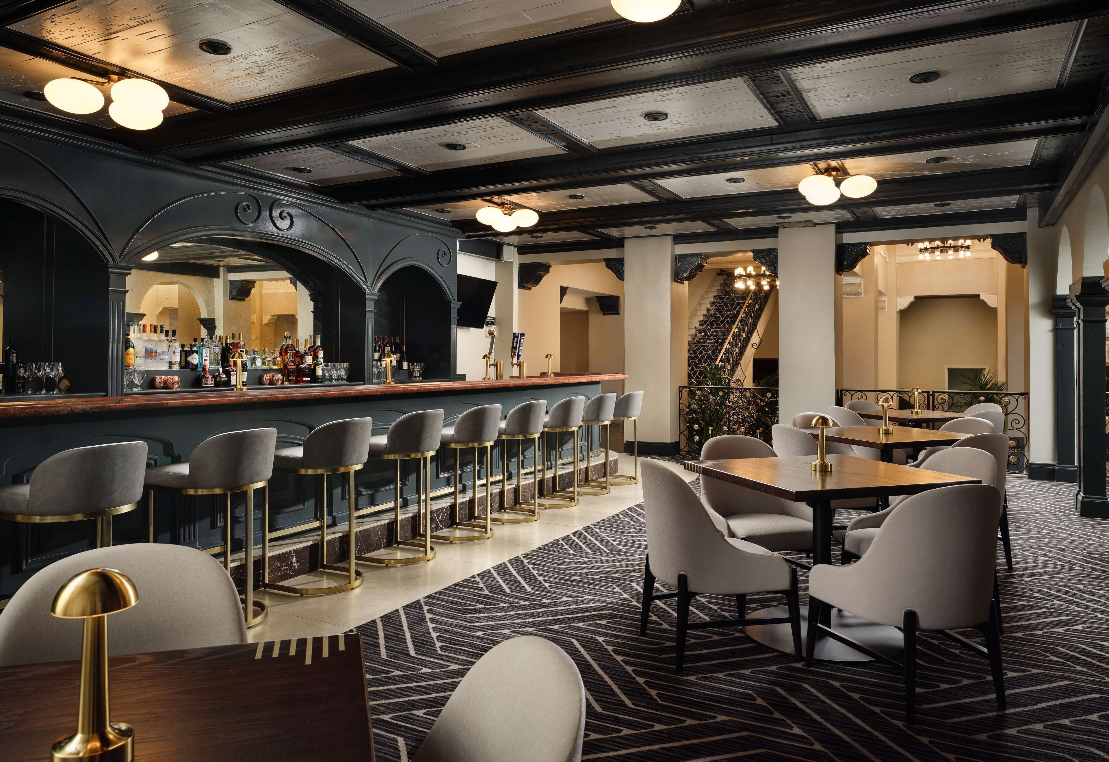 Uma área de bar com bancos altos e um balcão comprido, uma sala de jantar com mesas e cadeiras, e uma escadaria ao fundo.