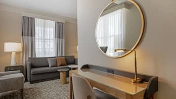 A hotel room with a sofa, a desk with a mirror, a lamp, and a view of the outdoors.