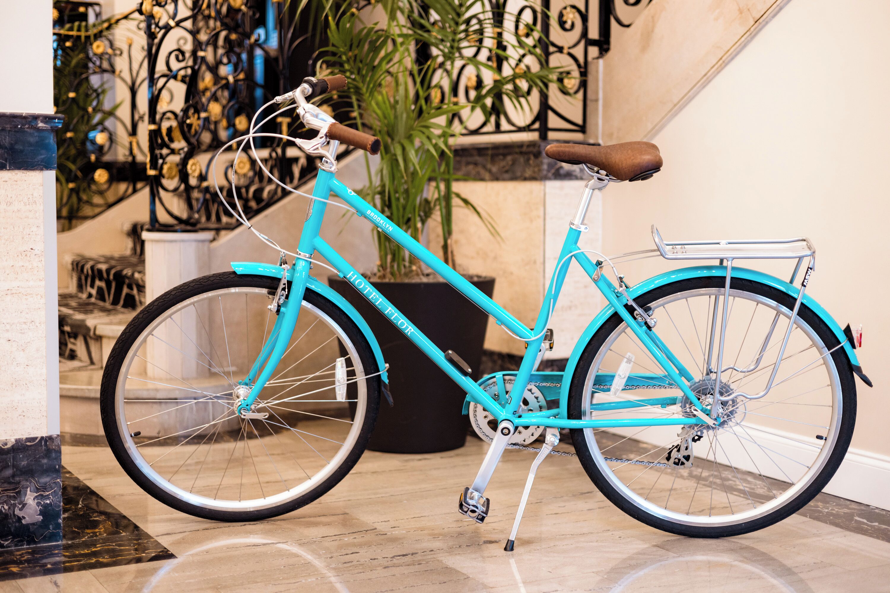 A turquoise bicycle with a brown seat and a rear rack, parked indoors near a staircase and a potted plant.
