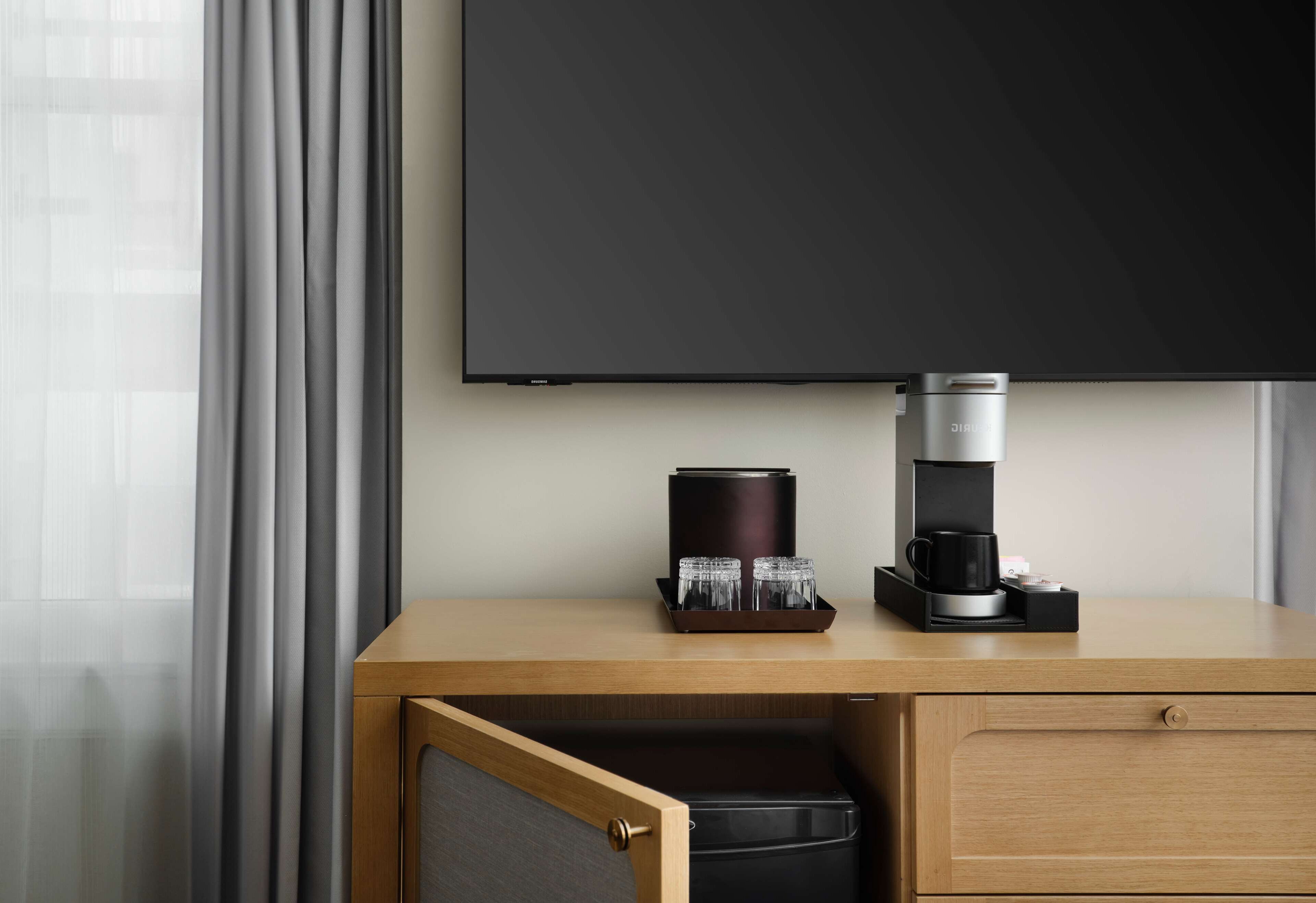 Ein Hotelzimmer mit einem Holztisch, einer Kaffeemaschine, einem Tablett mit Gläsern, einer schwarzen Kaffeekanne und einem Fernseher an der Wand.