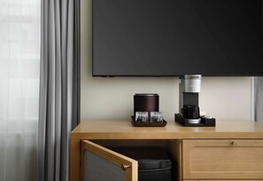 Une chambre d’hôtel avec un bureau en bois, une machine à café, un plateau avec des verres, une cafetière noire et un téléviseur fixé au mur.