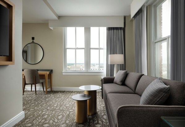 Habitación de hotel con un sofá gris, un espejo redondo, una mesita, un escritorio y un ventanal grande con cortinas.
