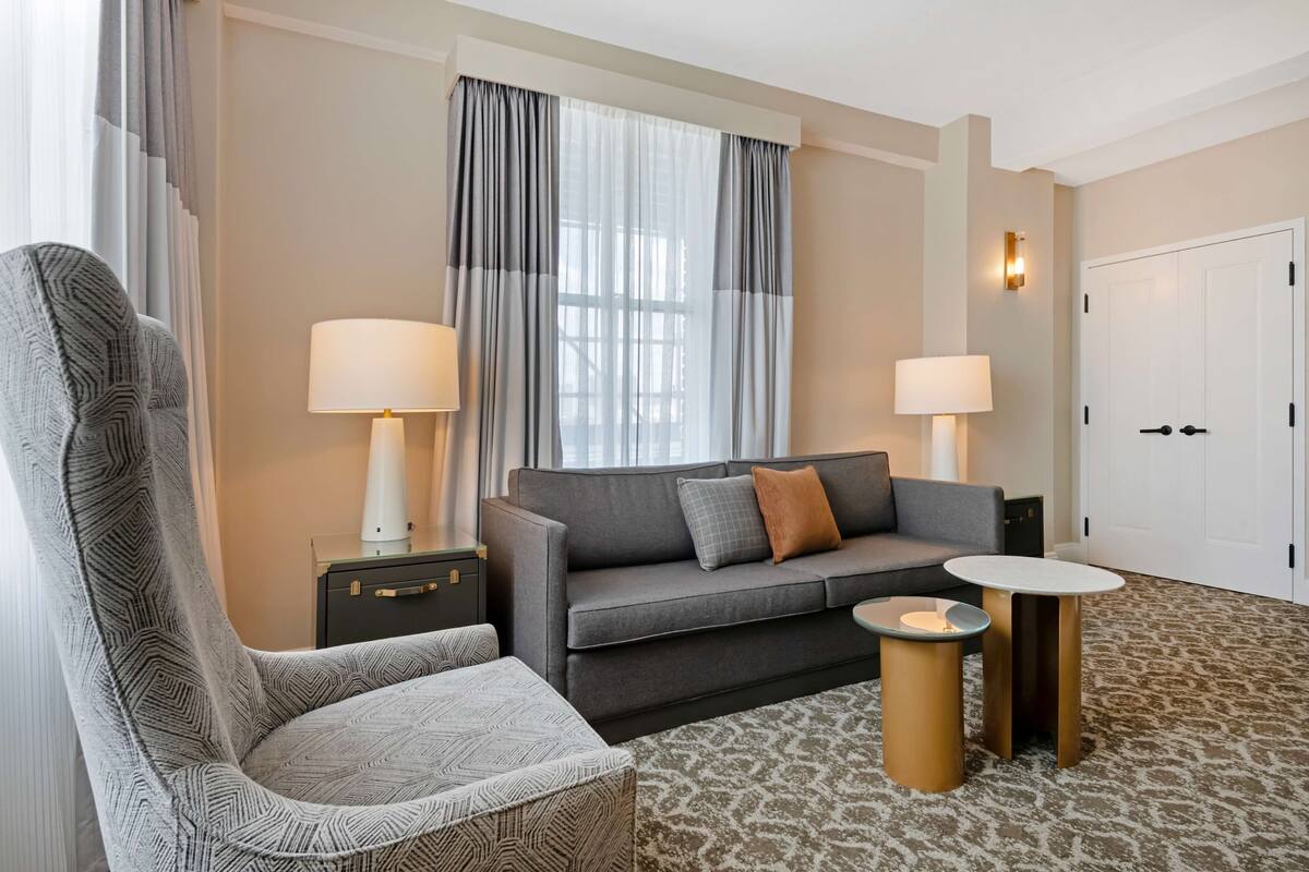 Een moderne hotelkamer met een grijze bank, een gestreept fauteuil, twee nachtkastjes met lampen en een kamerbreed tapijt met dessin.