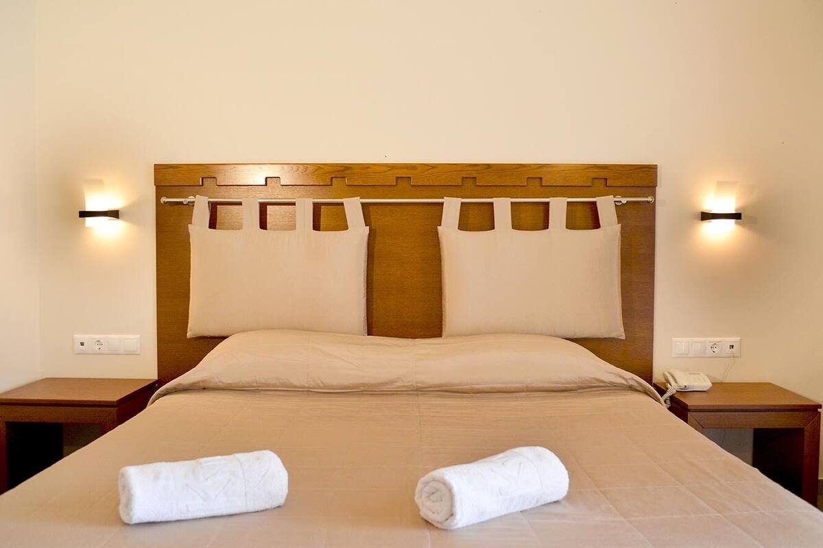 Una cama bien hecha con dos toallas enrolladas, un cabecero de madera y lámparas de noche.