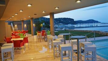 An outdoor seating area with tables and chairs, overlooking a body of water and a hilly landscape.
