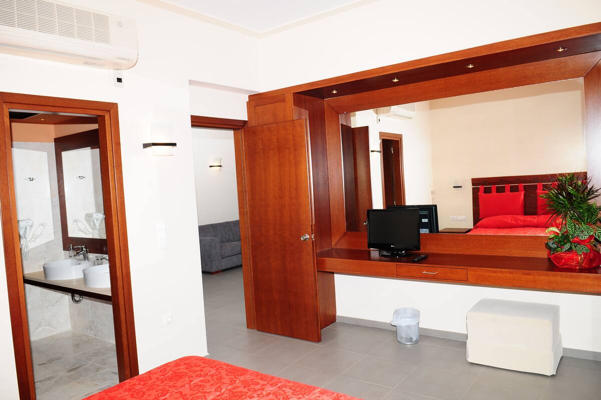 Una habitación de hotel moderna con un escritorio de madera, una computadora, una cama roja y un baño con espejo y lavamanos.