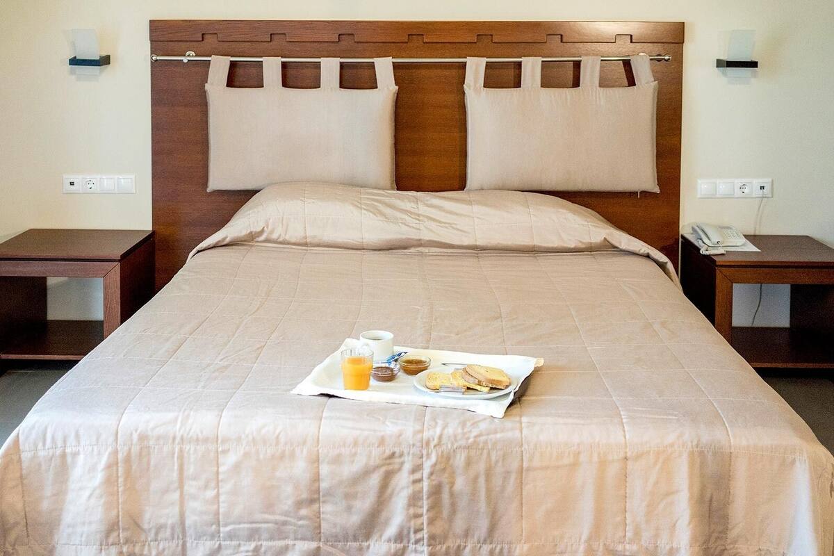 Una cama bien tendida con una bandeja de desayuno que incluye tostadas, una taza de café y un vaso de zumo de naranja.
