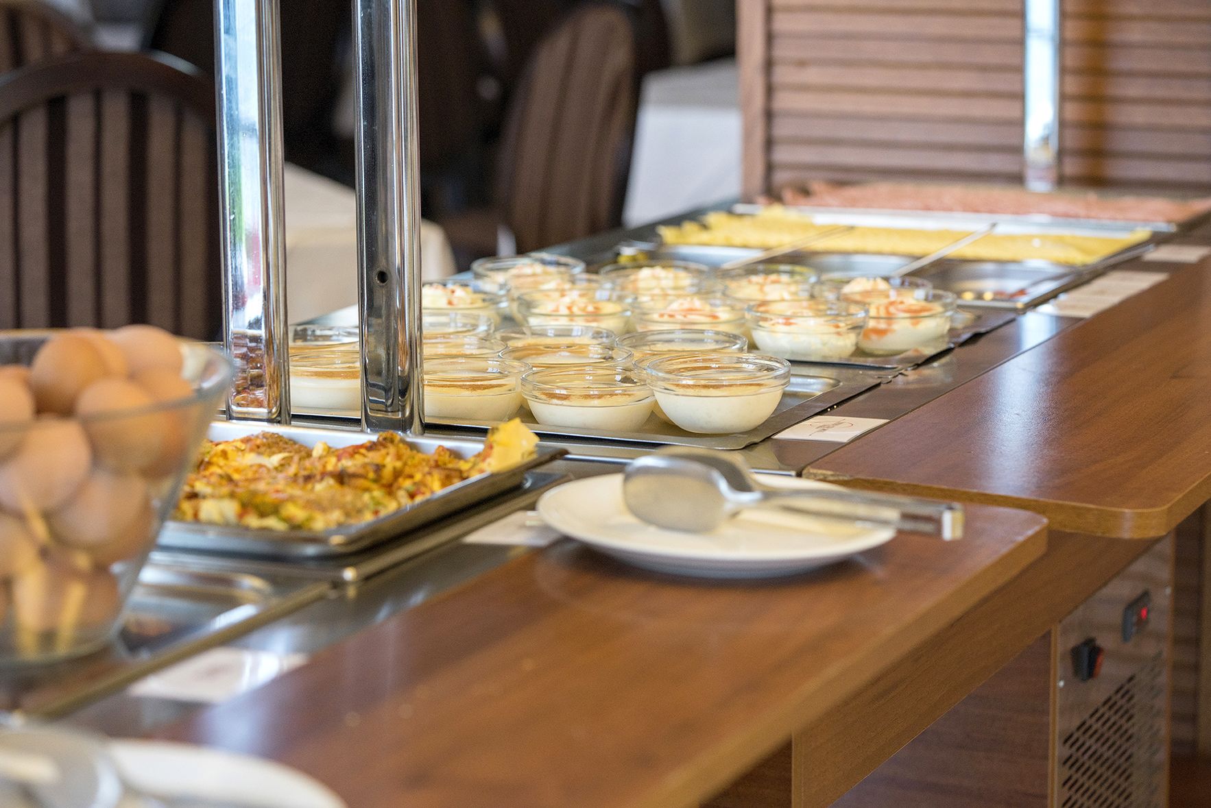 A buffet table with various food items, including a tray of scrambled eggs, bowls of yogurt, and a dish with a golden-brown crust.