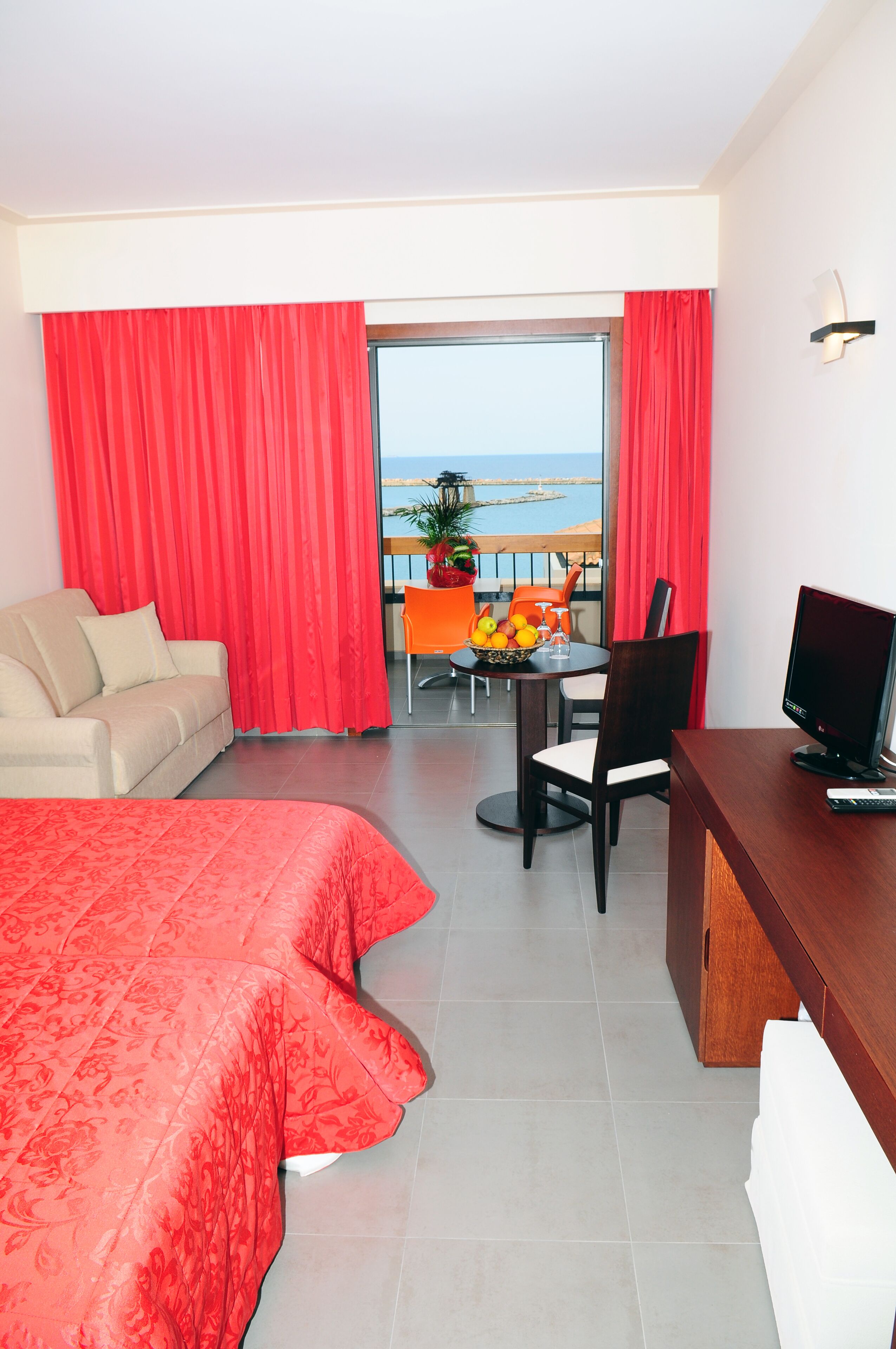 Habitación de hotel con cama, escritorio, televisor, balcón con mesa y sillas, y vista al mar.