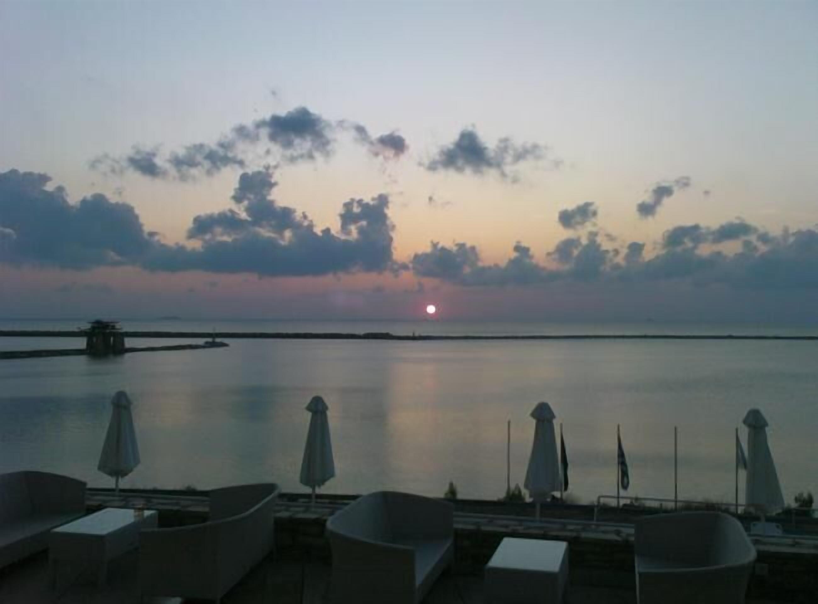 A sunset view from a terrace with lounge chairs and umbrellas, overlooking calm water and distant land.
