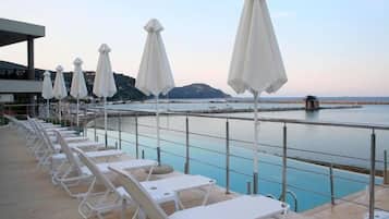 Uma área à beira da piscina com espreguiçadeiras brancas e guarda-sóis, vista desobstruída para o mar e um litoral rochoso ao fundo.
