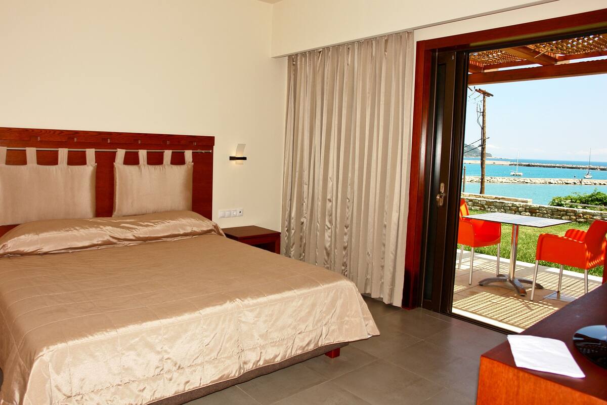 Habitación de hotel con cama, mesita de noche, escritorio y vista al mar a través de una puerta de vidrio.