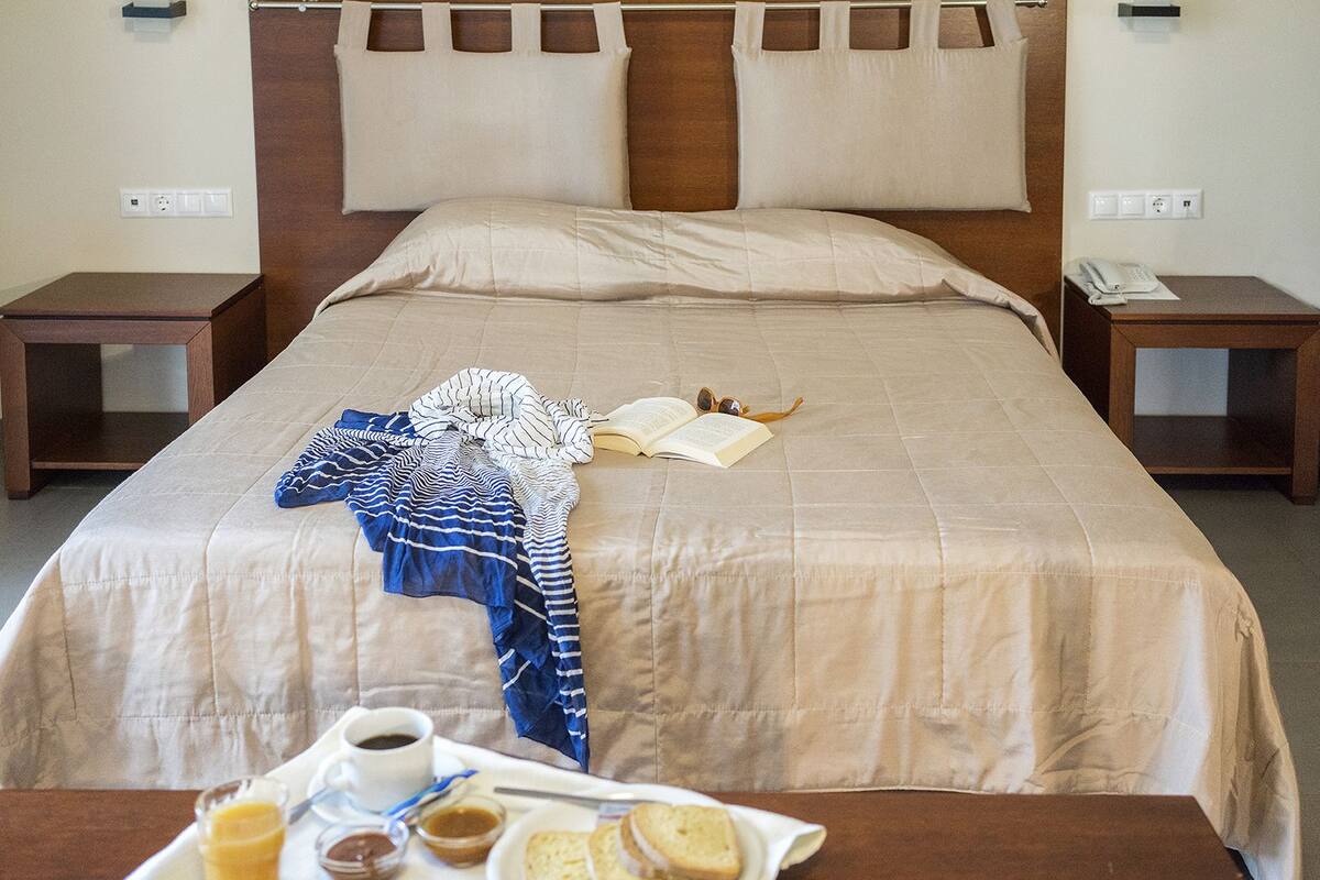Una cama bien hecha con edredón color beige, almohadas y una manta a rayas. Hay una mesita de noche con un libro, gafas y un teléfono. Un carrito con alimentos para el desayuno está en el suelo.