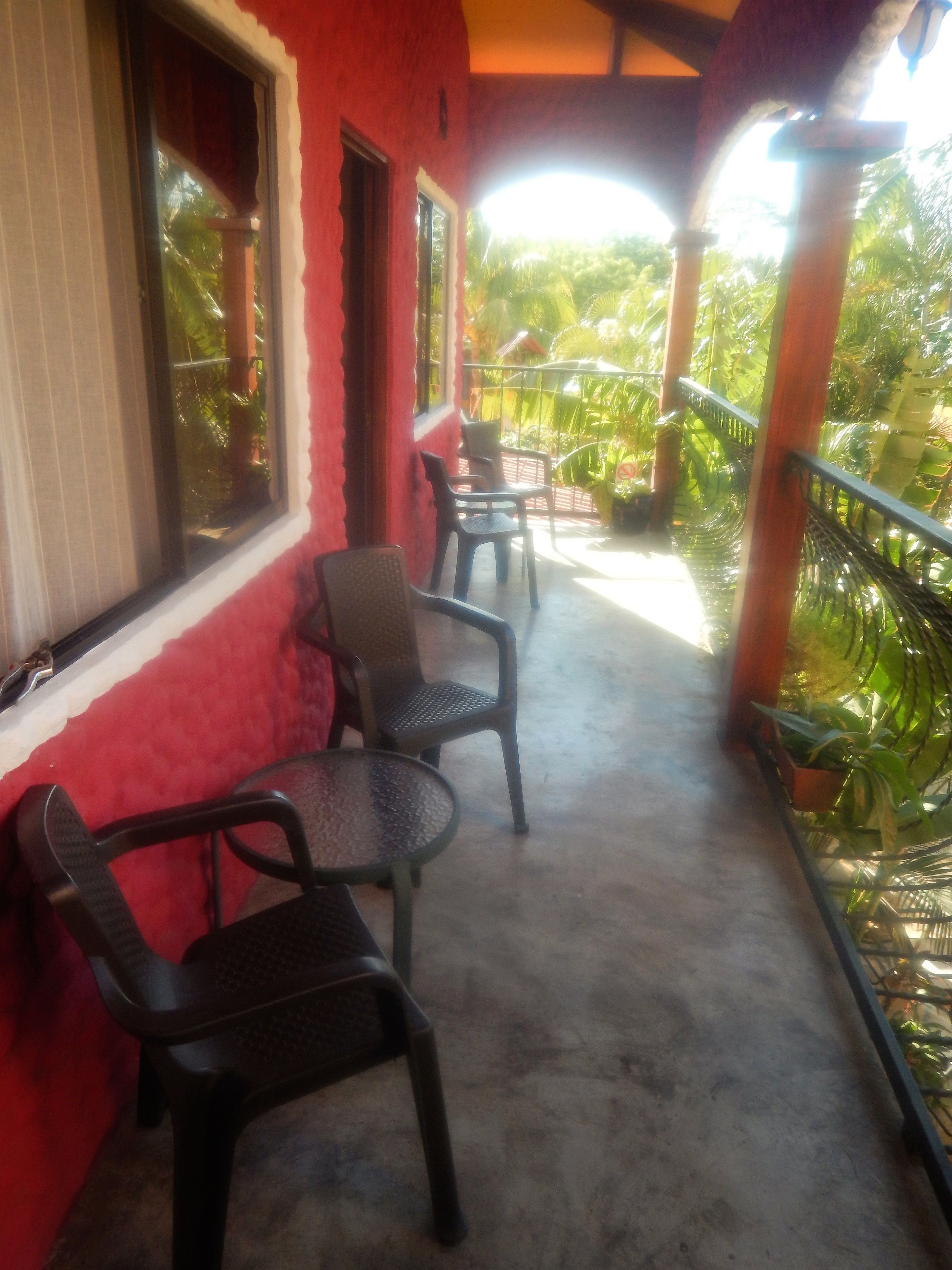 A covered outdoor seating area with chairs and tables, a red wall, and a view of greenery.