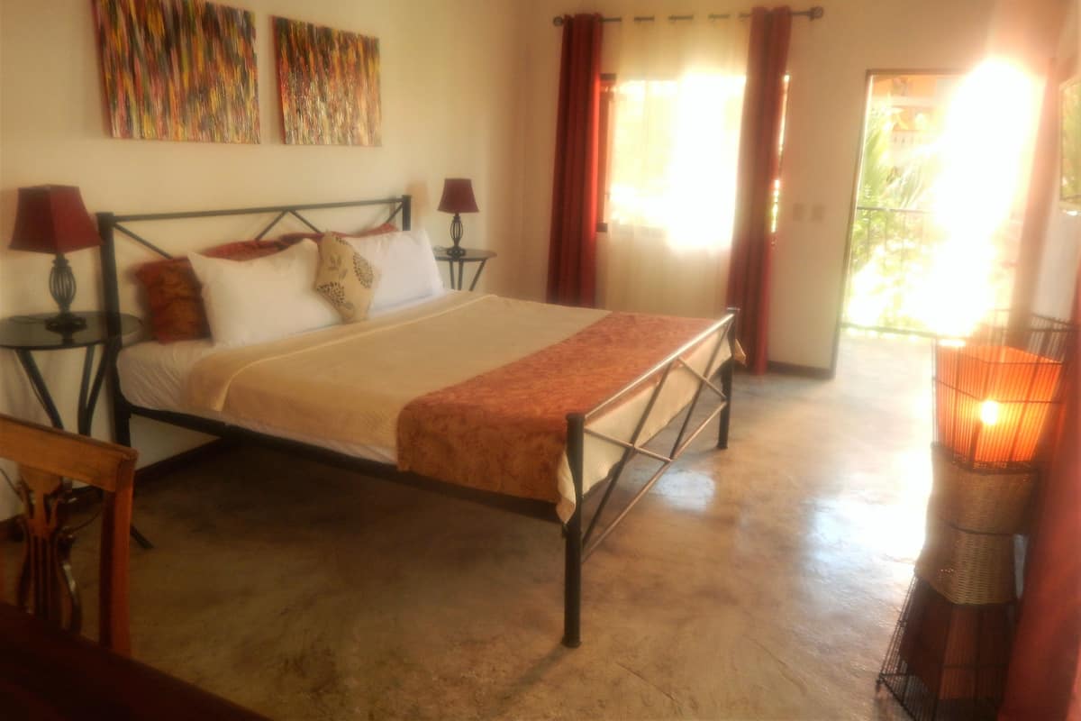 A bedroom with a bed, two bedside tables, a lamp, a window with curtains, and a painting on the wall.