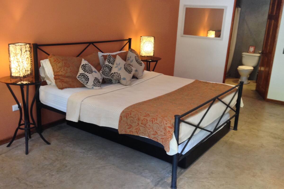 A bedroom with a bed, two bedside lamps, a bedside table, a mirror, and a toilet in the adjacent room.