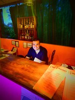 Una zona de bar con mostrador de madera, un atril para el menú y una estantería con botellas.