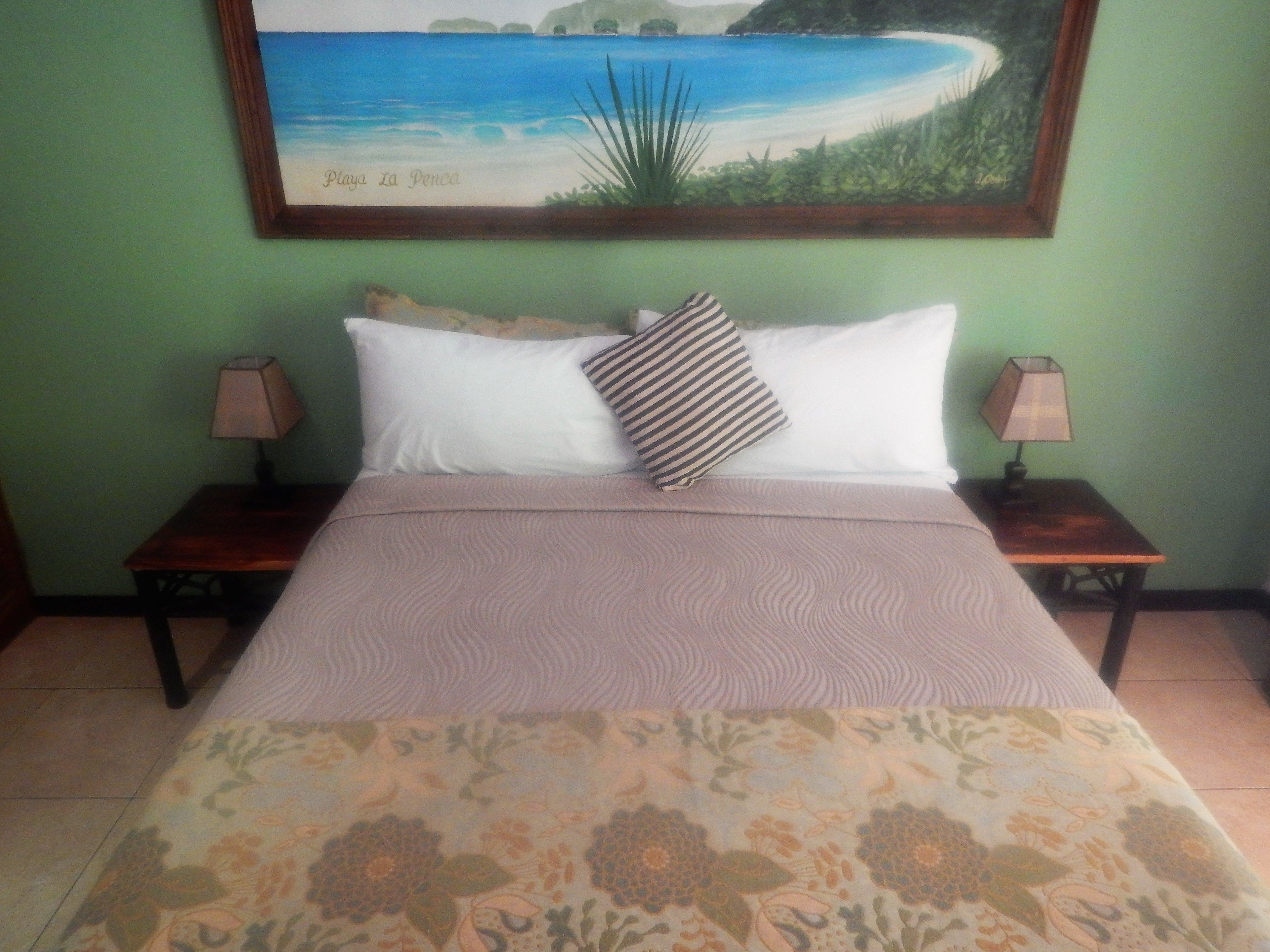 A bed with a floral bedspread, two bedside tables with lamps, and a painting of a beach scene on the wall.