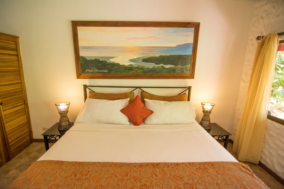 A bedroom with a bed, two bedside tables with lamps, a painting of a landscape, and a window with curtains.