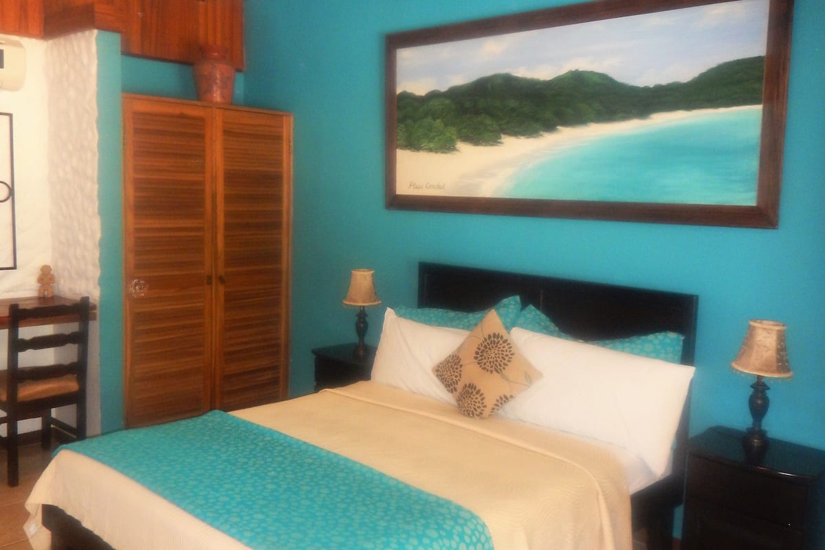 A bedroom with a bed, two nightstands, a wooden wardrobe, and a painting of a beach scene.