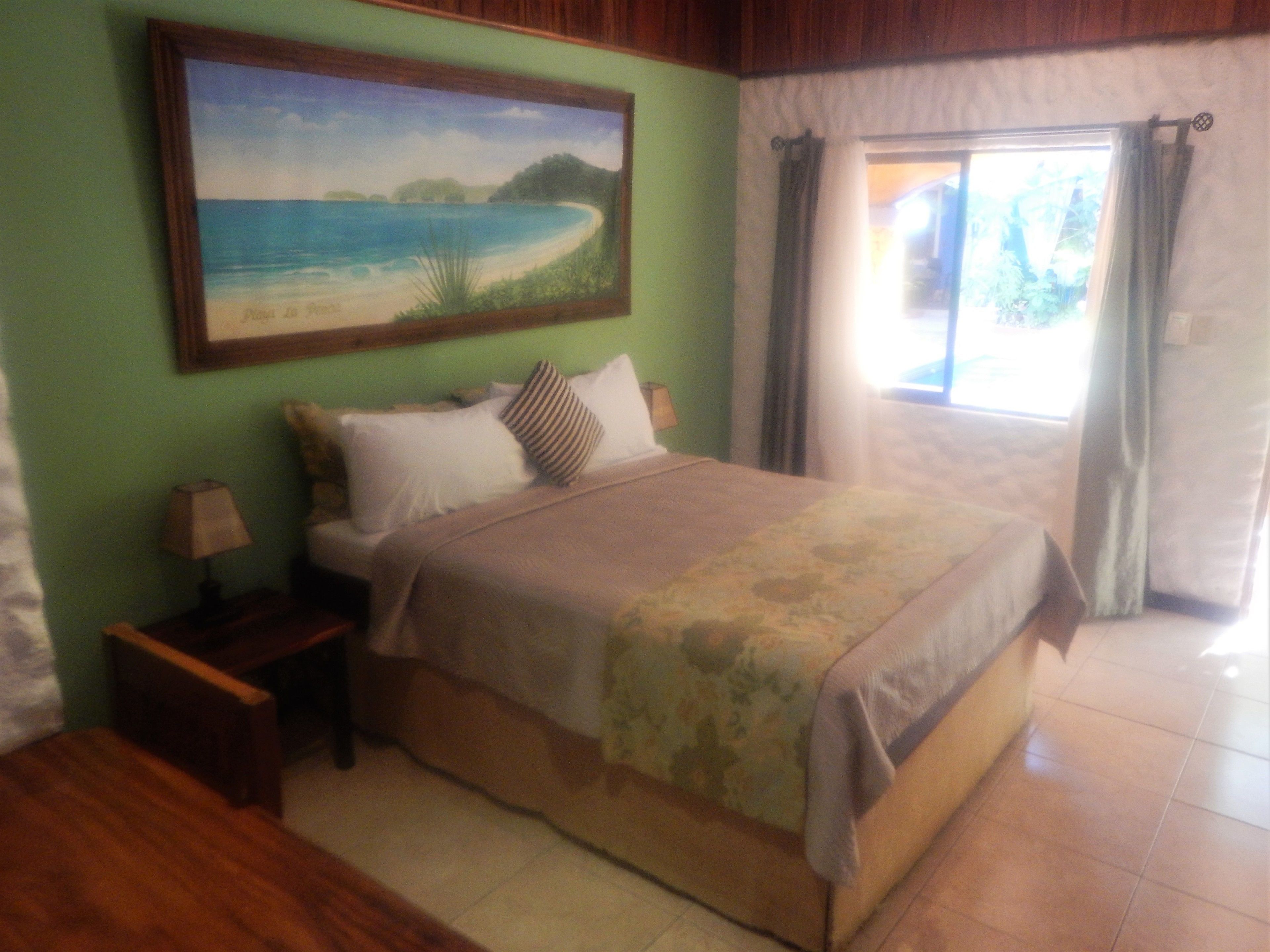 A bedroom with a bed, a nightstand, a lamp, a painting of a beach scene, and a view of the outdoors through a window.