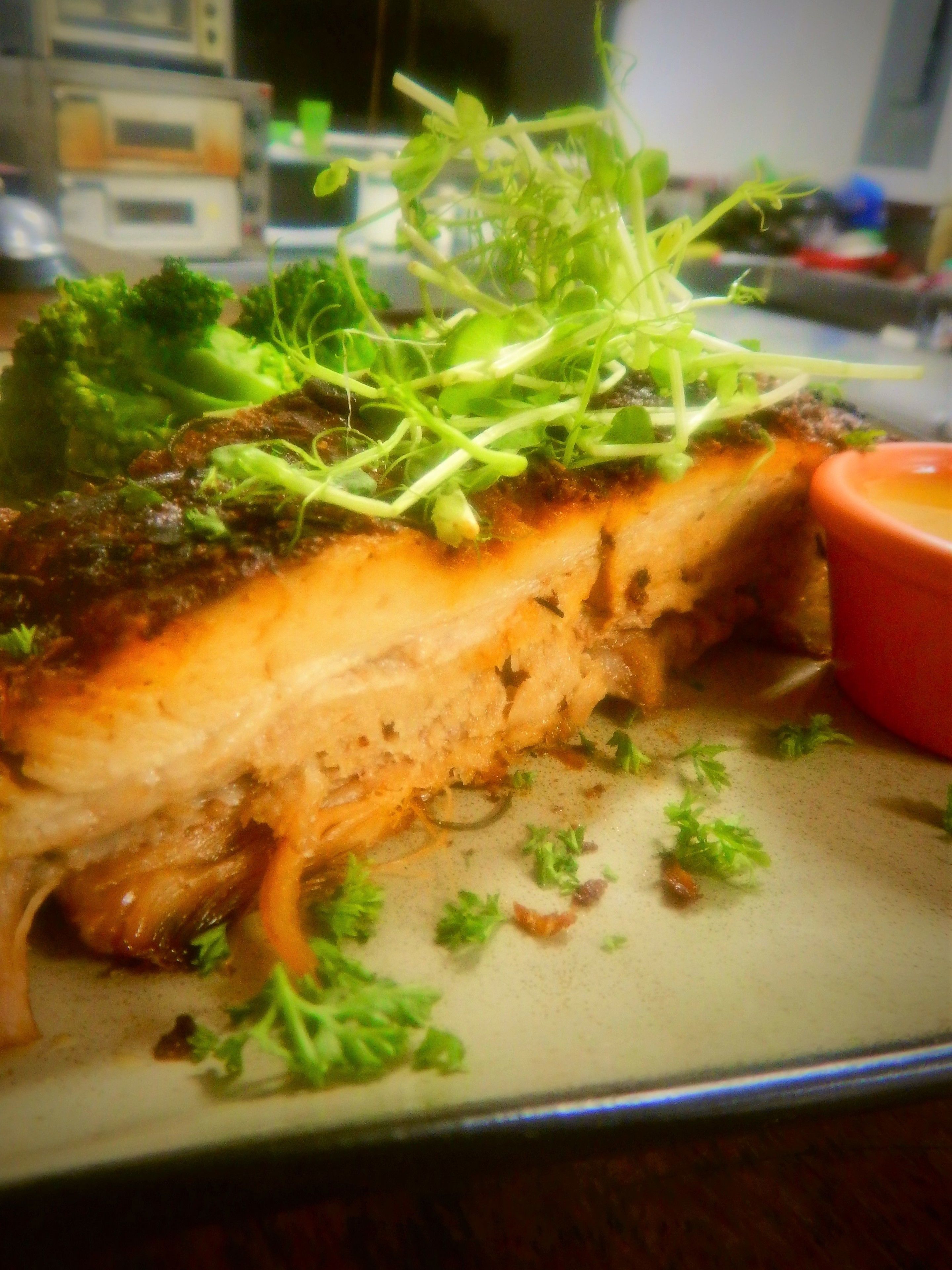 Un plato con un trozo de pescado a la parrilla, cubierto con hierbas verdes y una salsa roja pequeña.