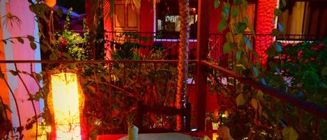 Un coin repas avec une table dressée pour deux personnes, entouré de plantes et d’un mur éclairé en rouge.