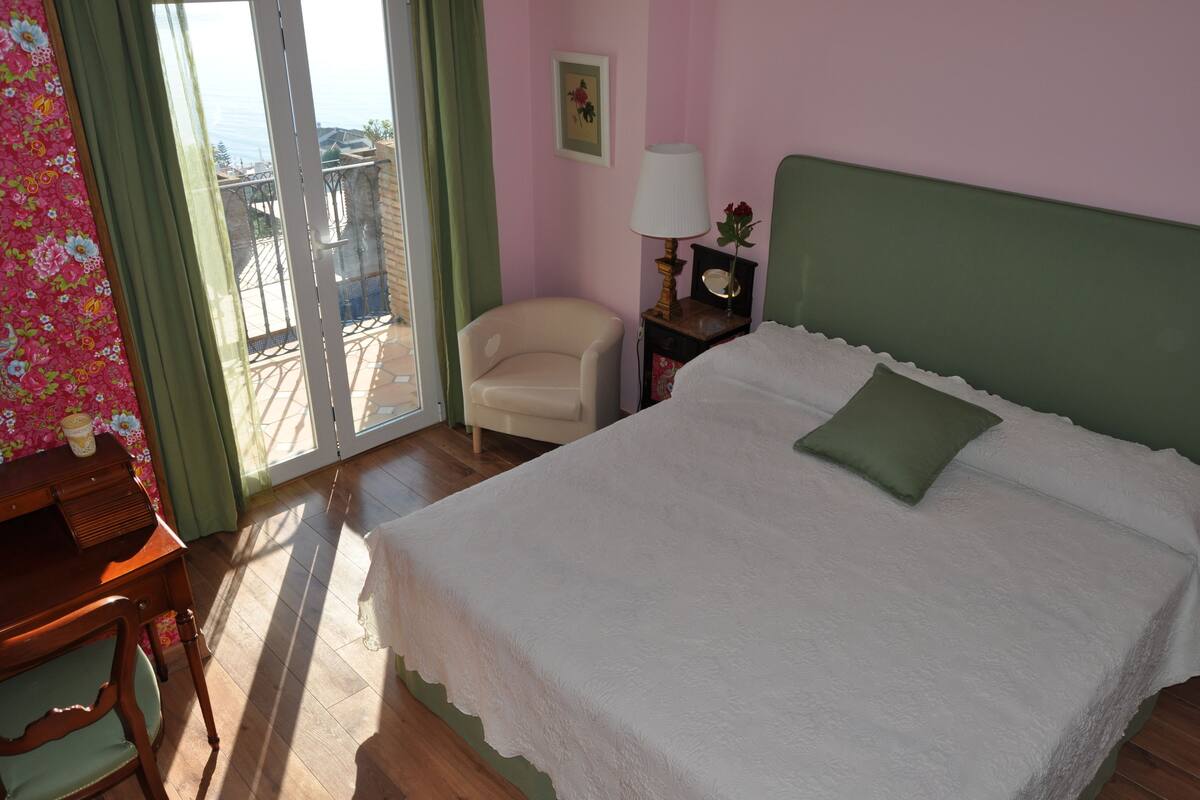 Un dormitorio con una cama, una silla, una mesita de noche, una lámpara y vistas a través de un ventanal corredero.