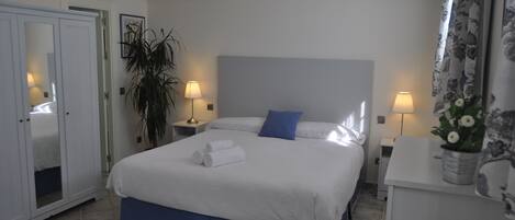A hotel room with a large bed, two bedside tables, a nightstand, a wardrobe, and a potted plant.