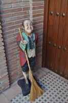 Une statue représentant un homme tenant un balai, debout sur un sol carrelé, avec un mur de briques et une porte en bois en arrière-plan.