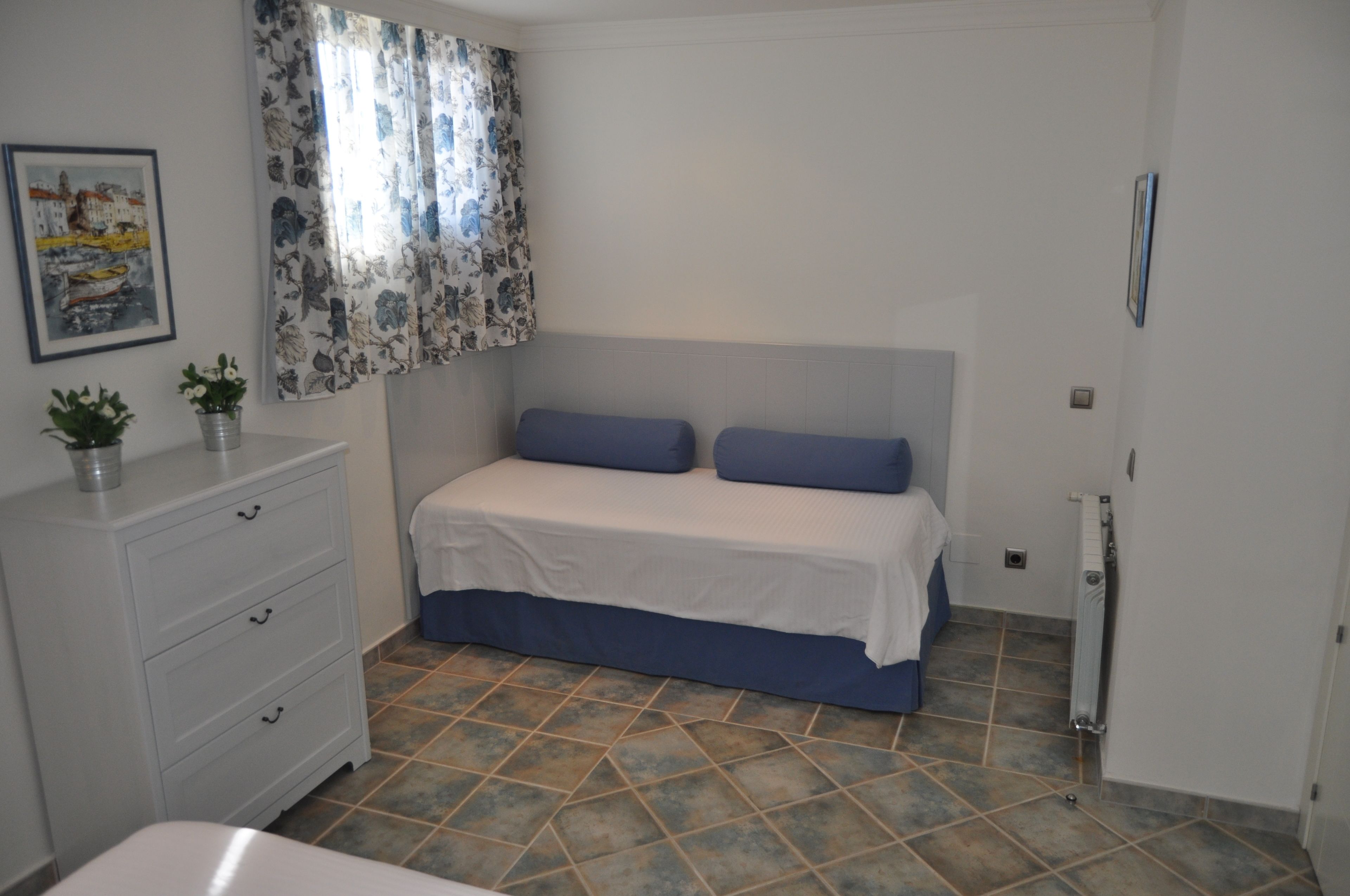Une chambre à coucher avec un lit, une commode, un radiateur et une fenêtre ornée de rideaux à motifs floraux.