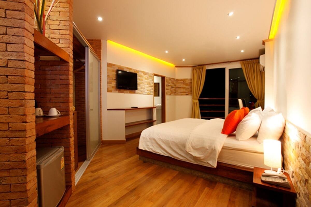 A modern bedroom with a brick accent wall, a flat-screen TV mounted above a desk, a bed with white bedding and orange pillows, and a nightstand with a lamp.