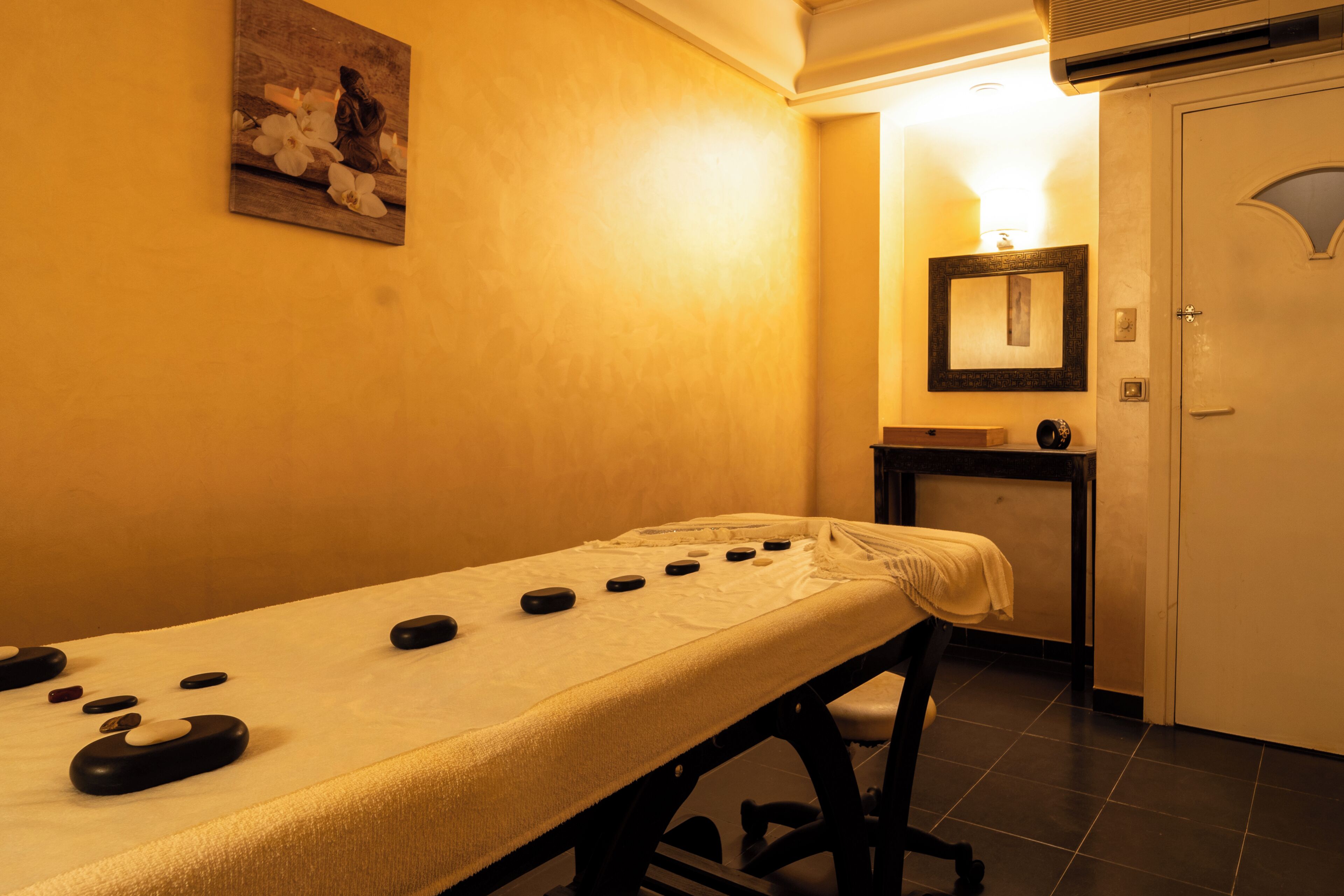 A massage table with stones, a small table with a lamp, and a door in a room with yellow walls.