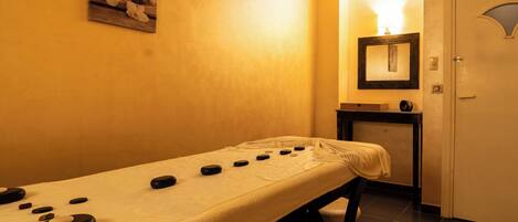 A massage table with stones, a small table with a lamp, and a door in a room with yellow walls.