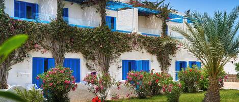 Un edificio blanco con contraventanas azules y un jardín con flores rosas y una palmera.
