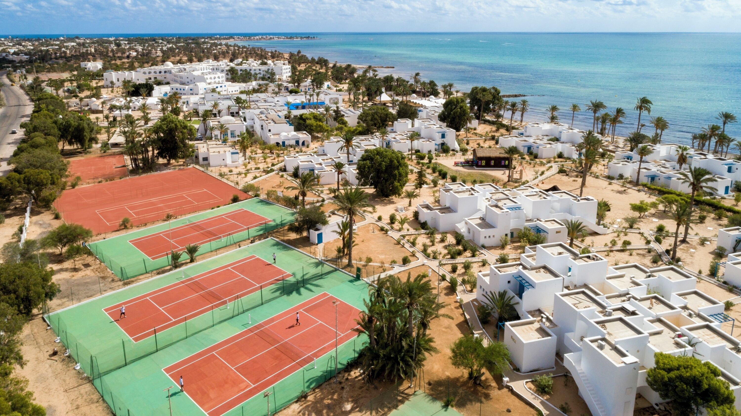 A coastal residential area with white buildings, a tennis court, and a beach in the background.