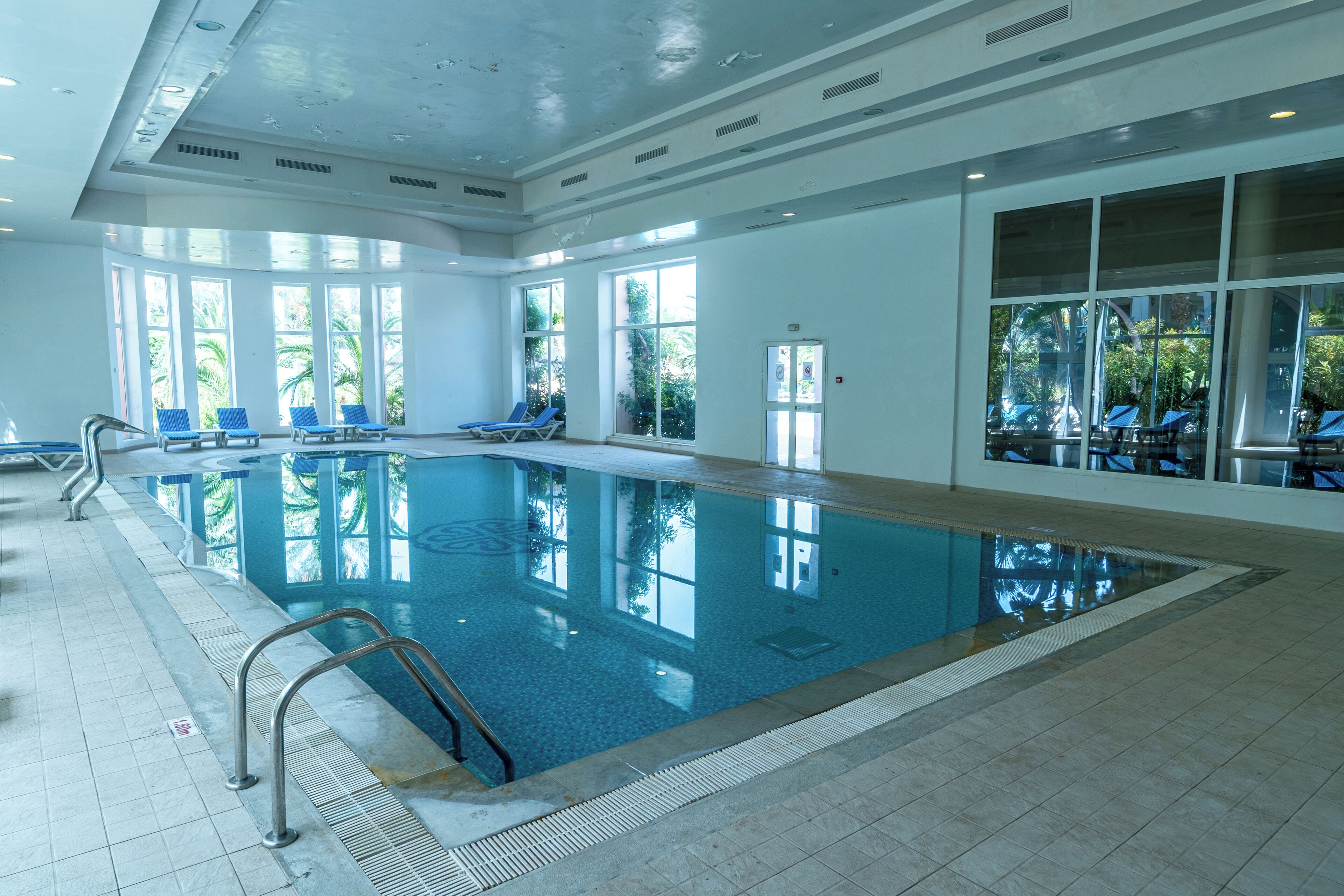 Ein Hallenbad mit klarem, blauem Wasser, umgeben von Liegestühlen und großen Fenstern.
