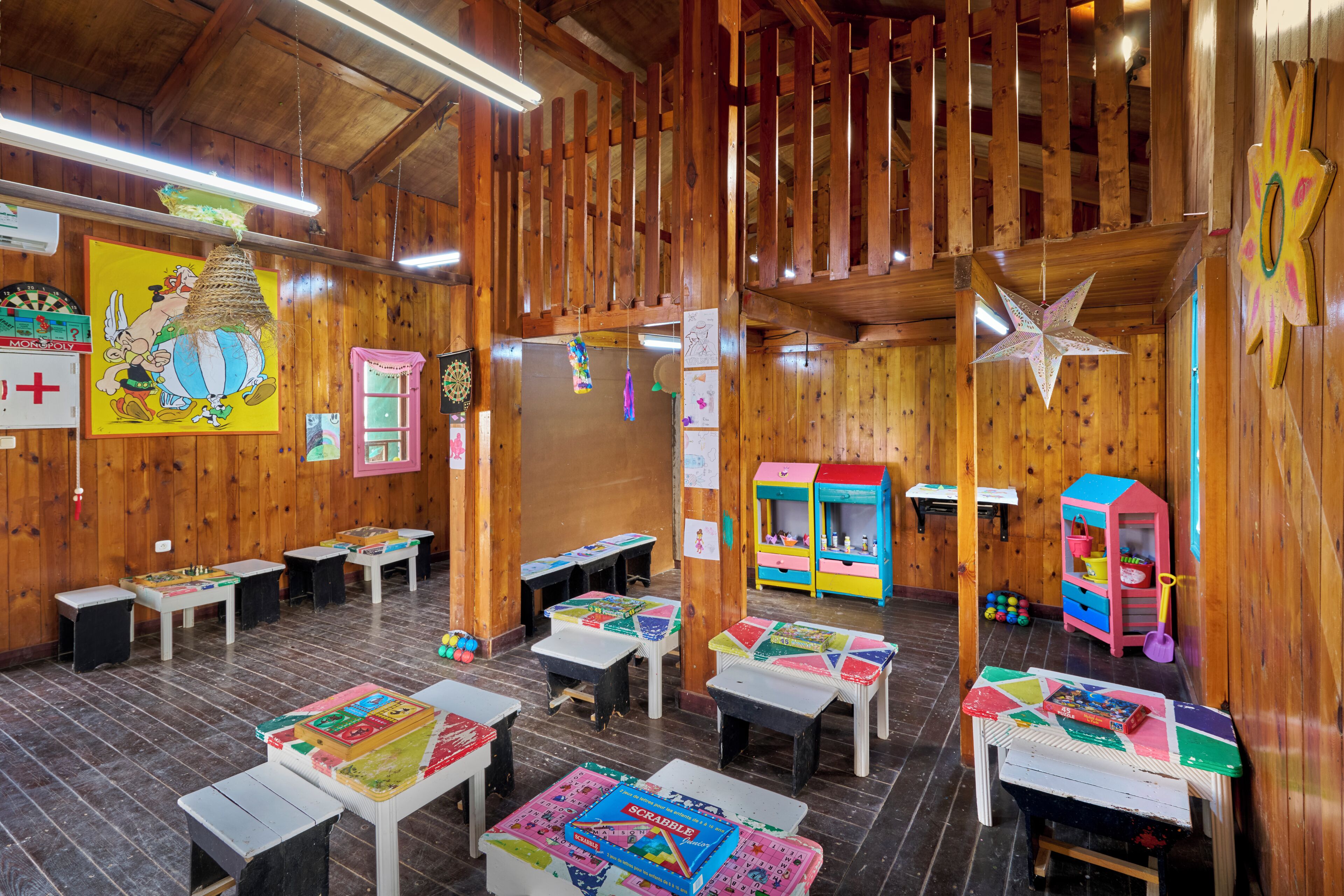 A room with wooden walls and ceiling, featuring tables with children's games, a colorful playhouse, and a star decoration.