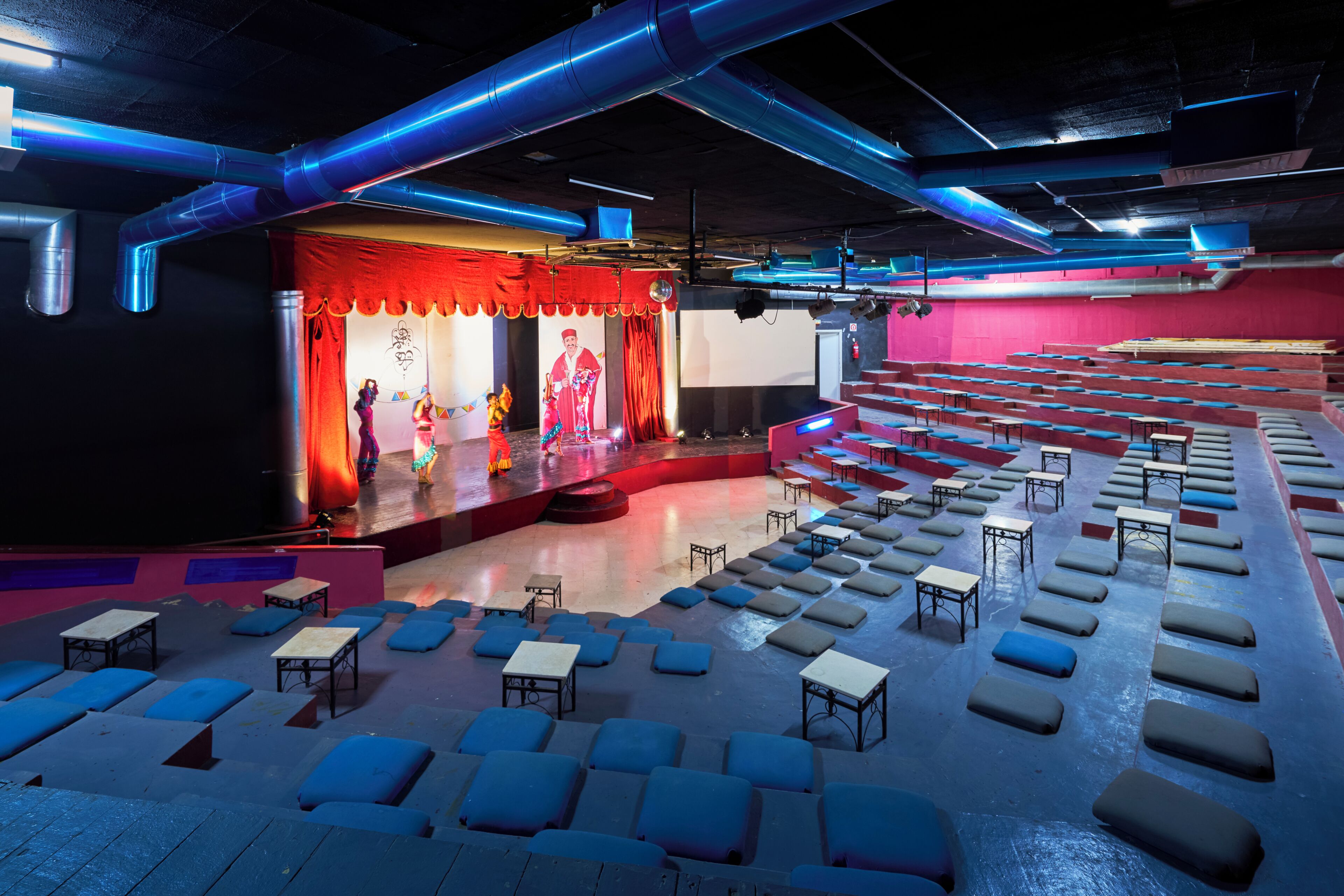 An auditorium with rows of blue cushioned seats, a stage with red curtains, and overhead blue pipes.
