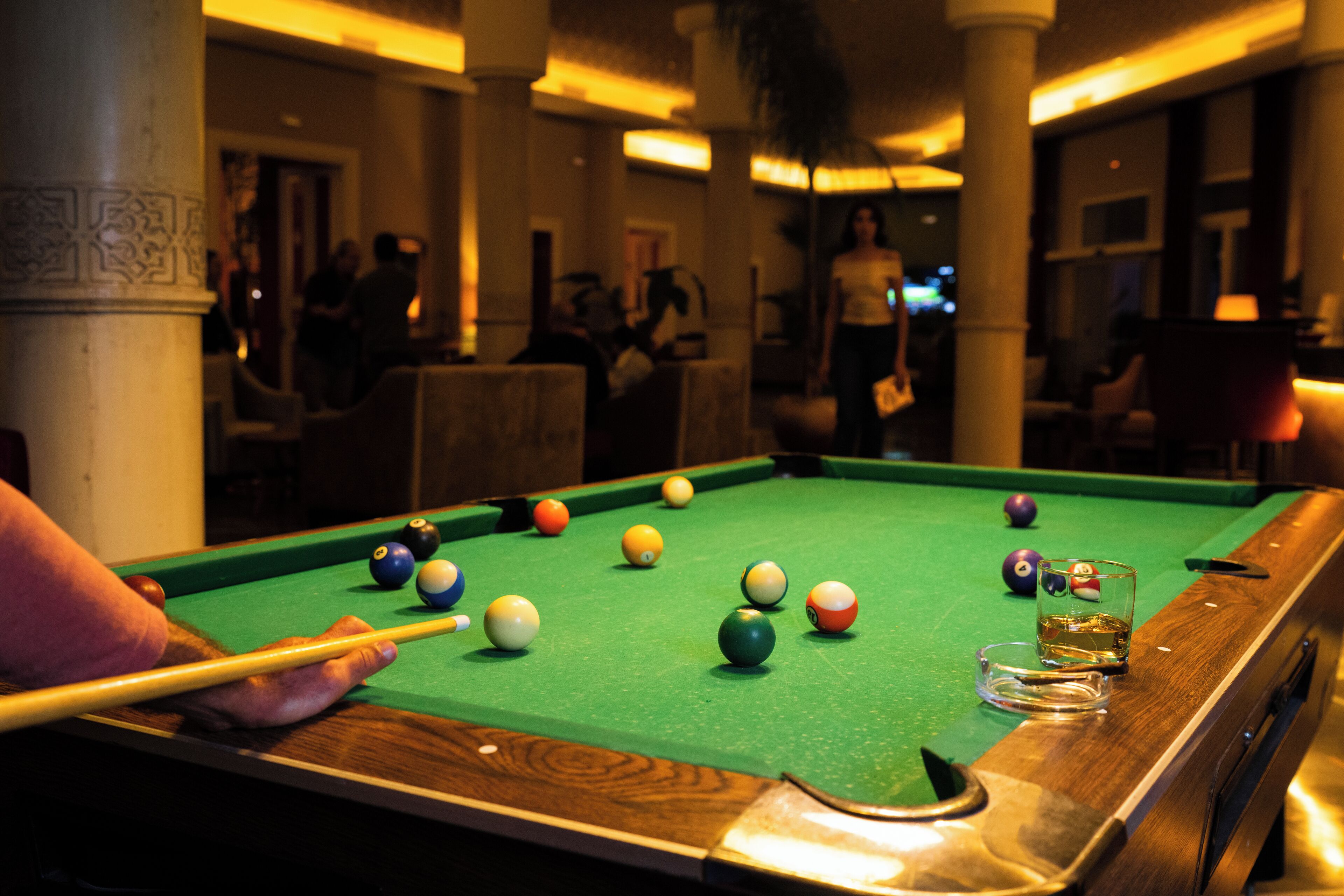 A pool table with balls and a cue stick, surrounded by chairs and a glass of beer.