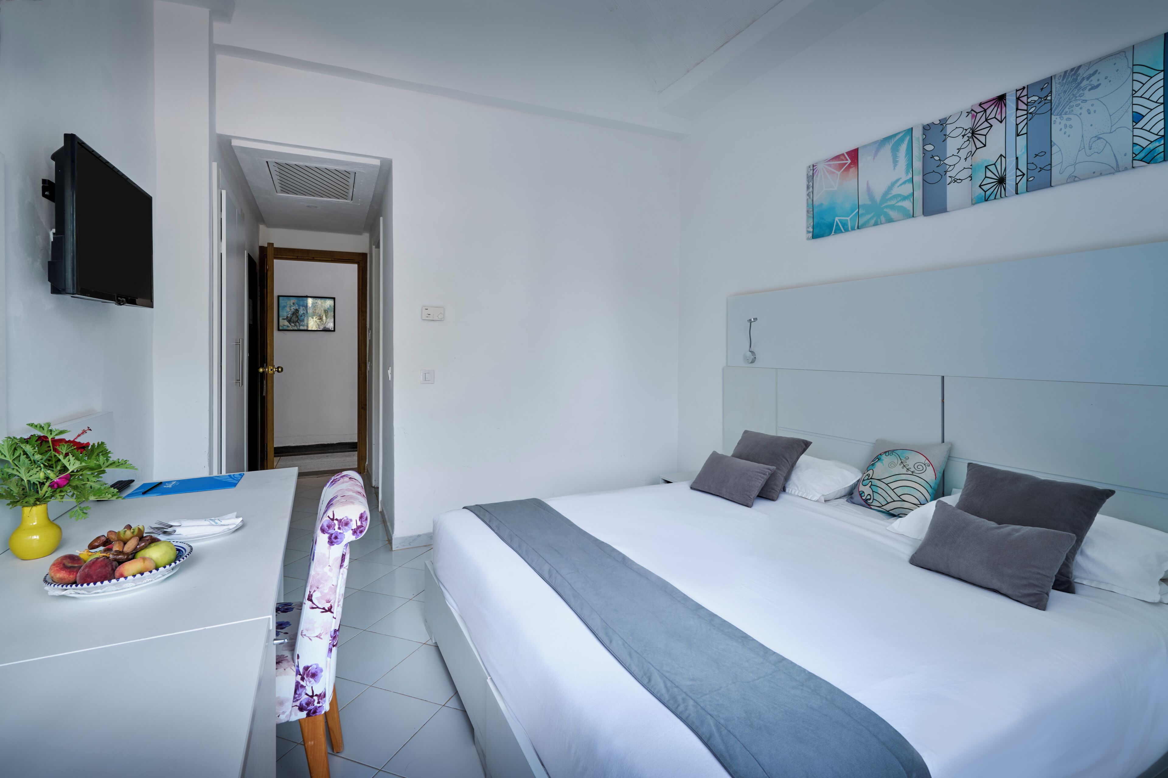 A hotel room with a bed, a desk with a vase of flowers, and a painting on the wall.