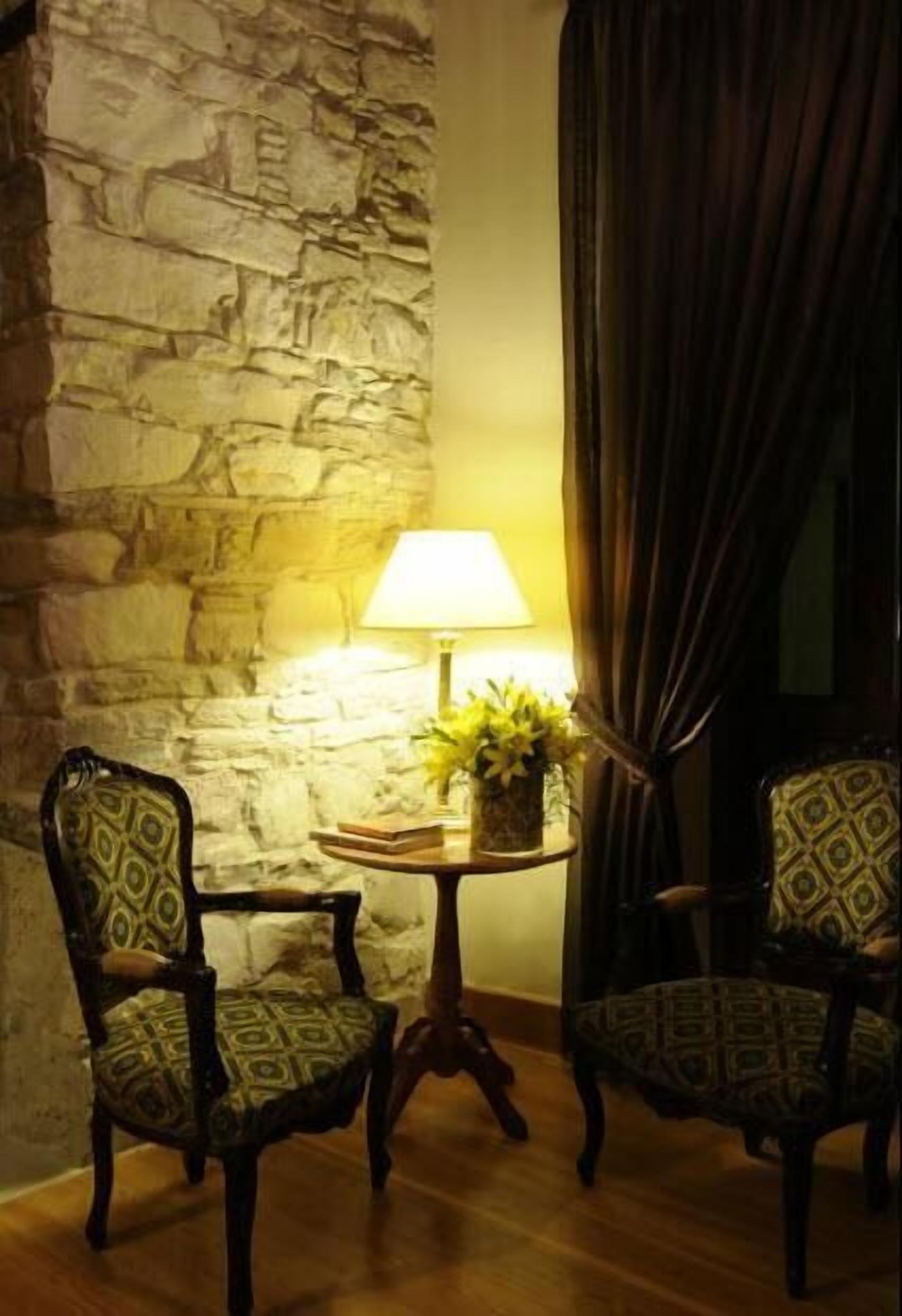 Una habitación con pared de piedra, dos sillas estampadas, una pequeña mesa redonda con una lámpara y flores, y una cortina oscura.
