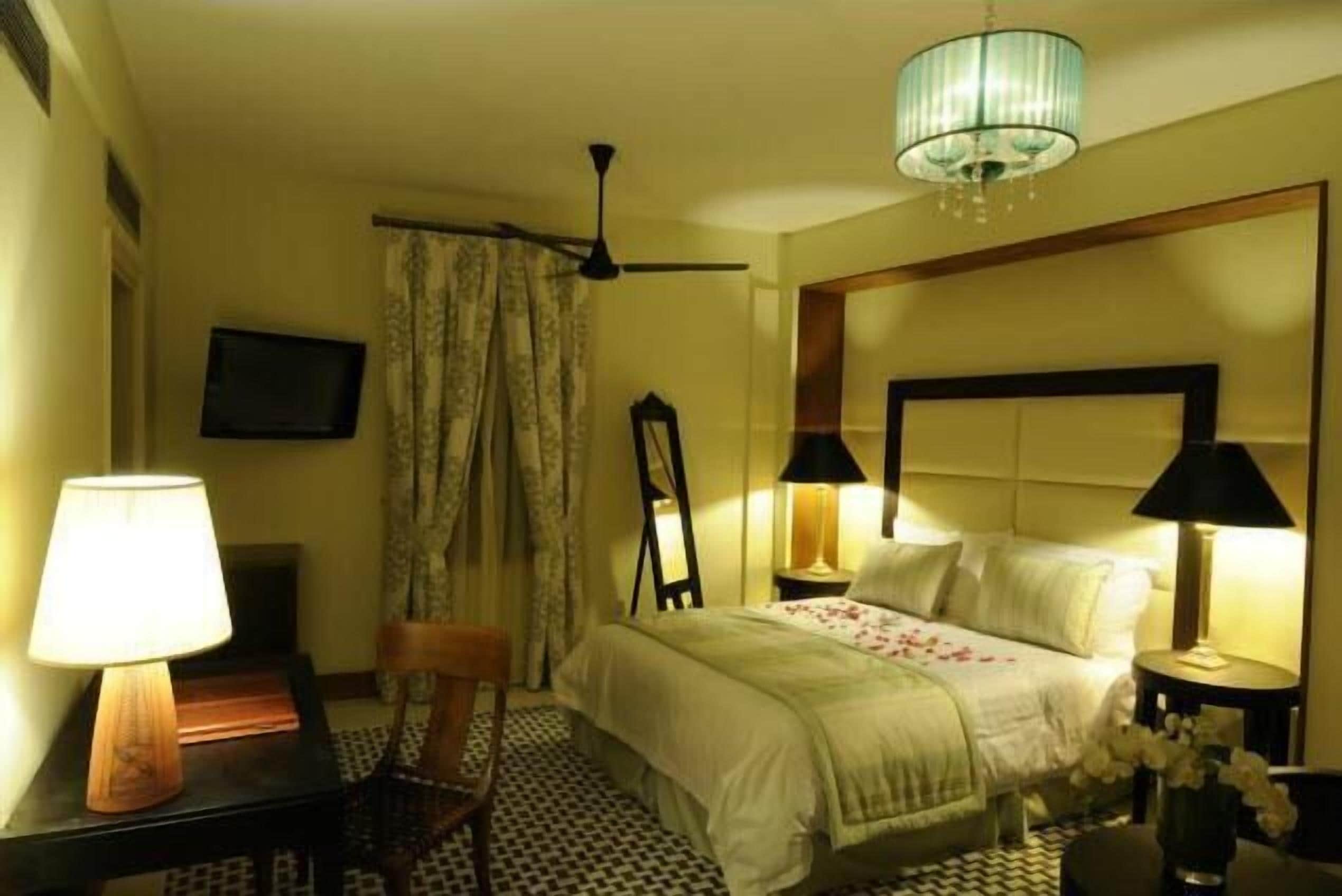 Habitación de hotel con cama, dos lámparas de noche, ventilador de techo, televisor y una silla.