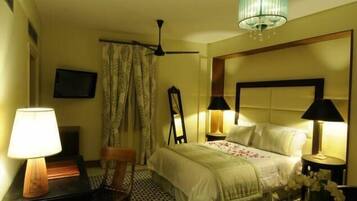 Habitación de hotel con cama, dos lámparas de noche, ventilador de techo, televisor y una silla.